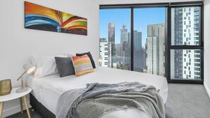 A modern bedroom with a city view, a bed with white linens, a colorful abstract painting, and a small bedside table with a lamp. - Riley - Resort Living * Gym Sauna Pool Wi-Fi (Southbank)