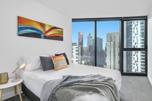 A modern bedroom with a city view, a bed with white linens, a colorful abstract painting, and a small bedside table with a lamp. - Riley - Resort Living * Gym Sauna Pool Wi-Fi (Southbank)