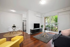 Ein modernes Wohnzimmer mit Flachbildfernseher, Essbereich mit Stühlen, Sofa und Balkon mit Grünblick.