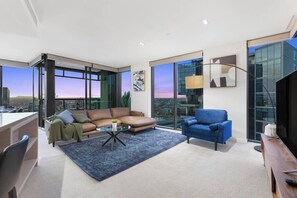 A modern living room with a sofa, armchair, and coffee table, large windows with a city view, and a glass partition. - Gia - Jaw-dropping 3BDR 2car executive apartment. (Docklands)