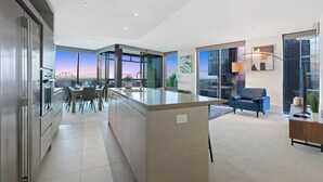 Eine moderne Küche mit einer zentralen Kochinsel, einem Essbereich mit Stühlen, einer Couch und großen Fenstern mit Blick auf die Stadt.