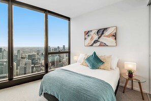 Ein Schlafzimmer mit großem Fenster, einem Bett mit blauer Decke, Blick auf die Stadt und einem Bild an der Wand.