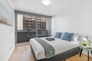 Ein modernes Schlafzimmer mit einem großen Bett, einem Fenster mit Blick auf Gebäude, einem kleinen Tisch mit Lampe und einer Pflanze.