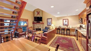 A spacious living area with a wooden staircase, a stone fireplace, a television, and a kitchen with bar stools. - Ski-in/ski-out condo w/ grill, fast WiFi, fireplace, shared pool/hot tub/laundry (Vail)