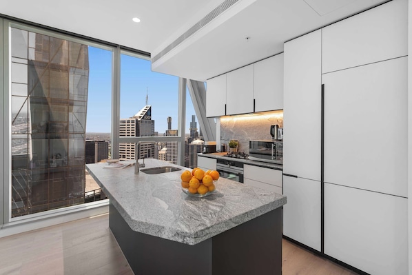 A modern kitchen with a large island, a bowl of oranges, and a city view through large windows. - Executive stunner in West Side Place with Parking (Melbourne)