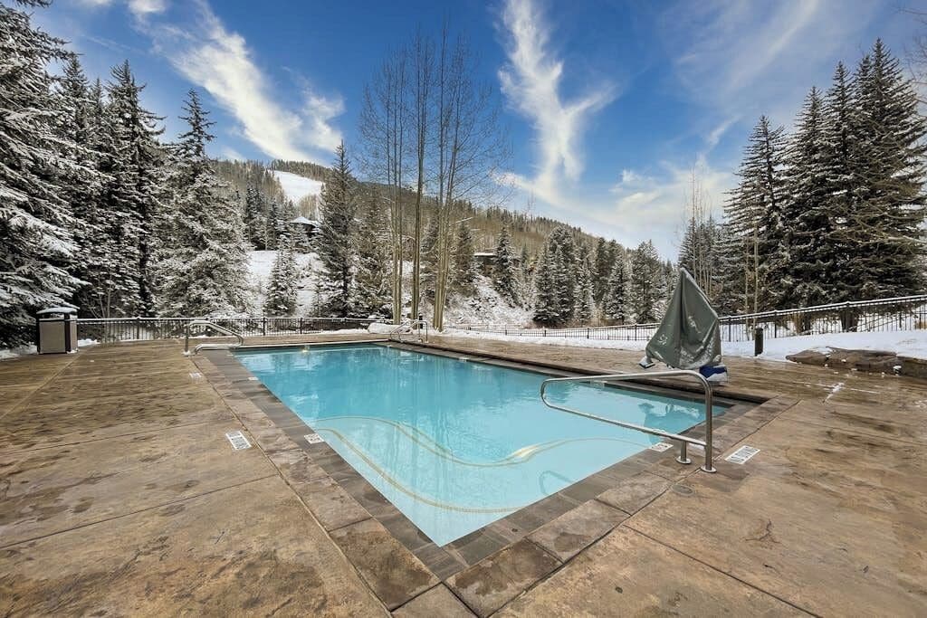 A swimming pool with a clear blue water surface, surrounded by a stone deck, and set against a backdrop of snow-covered trees and a clear sky.