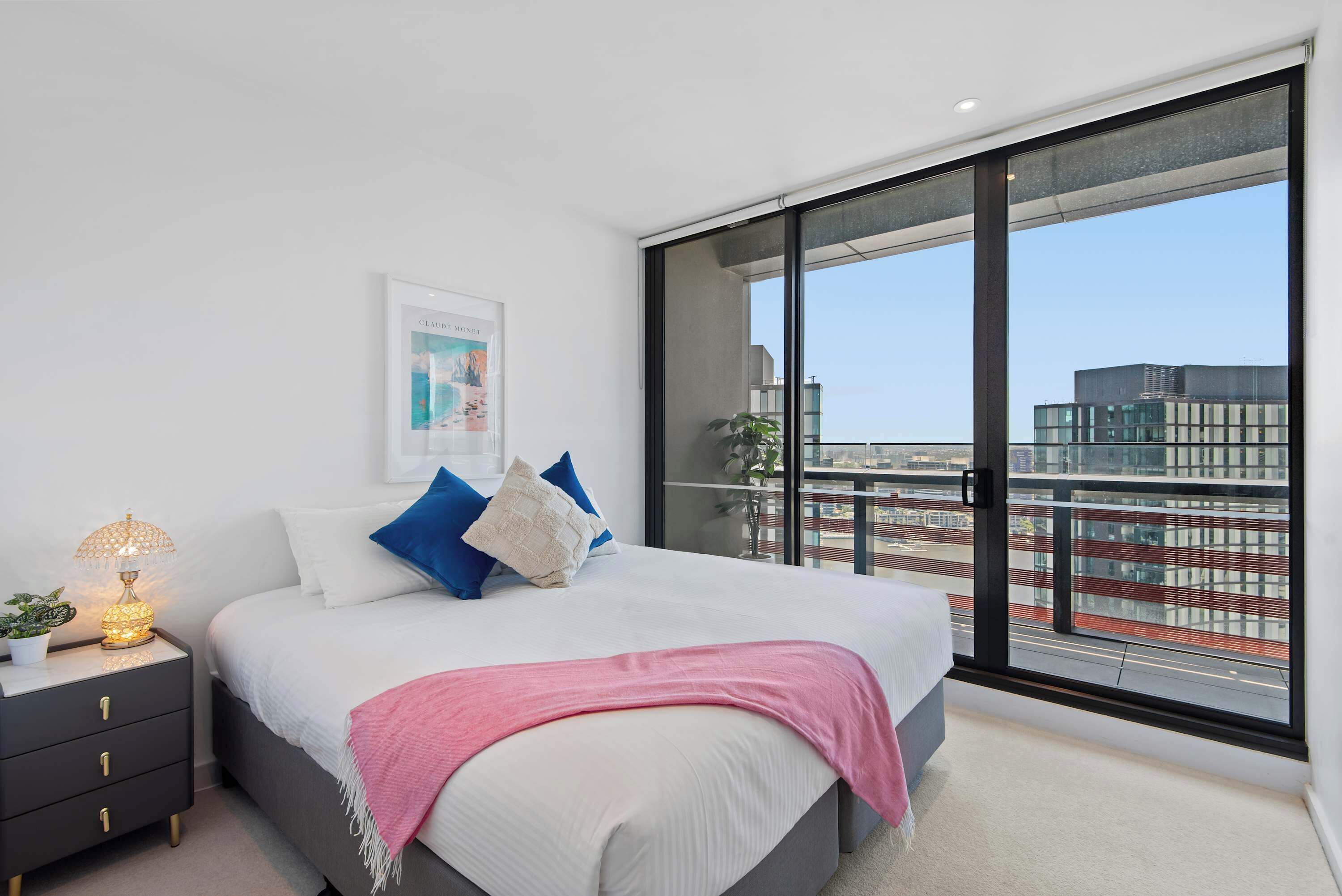 A modern bedroom with a large window, a bed with pillows, a bedside table with a lamp, and a framed picture on the wall.