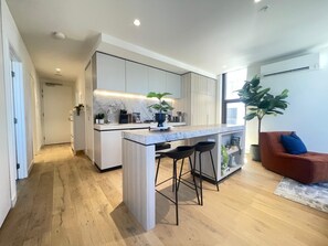 A modern kitchen with a central island, bar stools, and a seating area with a sofa and a large plant. - Southbank stunner with parking views gym and pool! (South Melbourne)