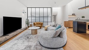 A modern living room with a flat-screen TV, a round coffee table, a sofa, and a kitchen area with a sink and countertop. - Architectural Masterclass in UberHip Northcote. (Northcote)