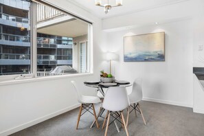 A modern dining area with a round table, white chairs, and a view of a building through large windows. - Stunning Large 2BD Parkside Apartment with Parking (Melbourne)