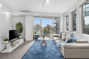 A modern living room with a flat-screen TV, a glass coffee table, a beige sofa, and a large window. - Stunning Large 2BD Parkside Apartment with Parking (Melbourne)