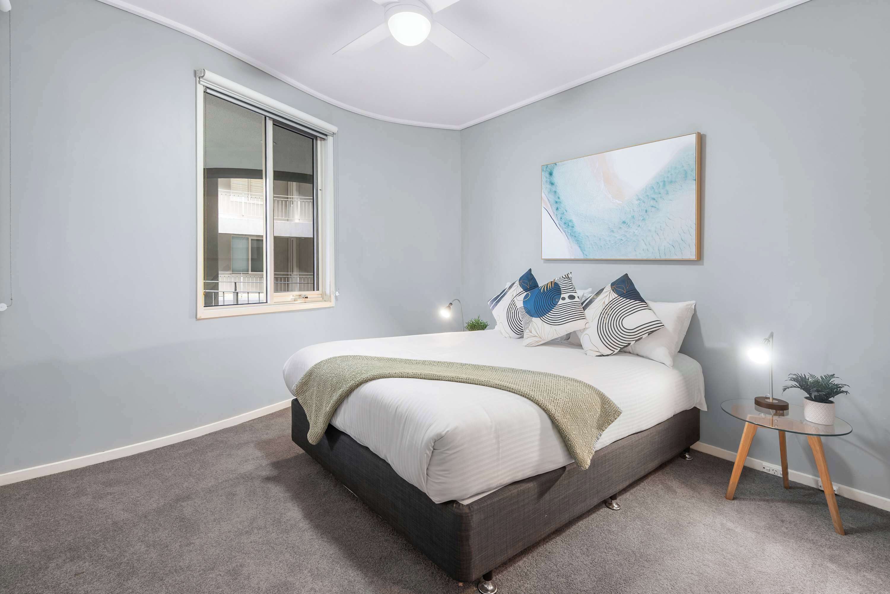 A bedroom with a bed, a window, a ceiling fan, a wall-mounted artwork, and a bedside table with a plant.
