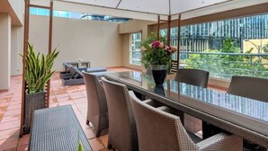 Un patio luminoso con un tavolo in vetro, sedie in vimini, piante in vaso e vista su un edificio.