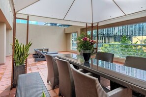 Eine sonnendurchflutete Terrasse mit einem Glastisch, Korbstühlen, Blumentöpfen und Blick auf ein Gebäude.