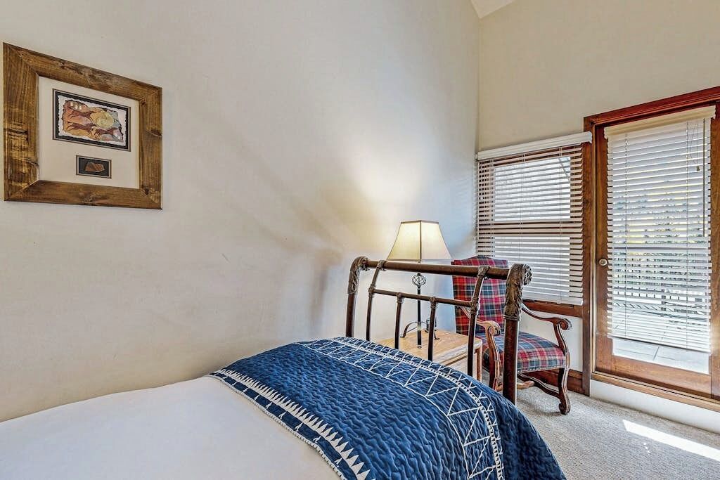 A bedroom with a bed, a chair, a lamp, a window with blinds, and a framed picture on the wall.