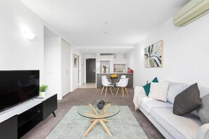 Ein modernes Wohnzimmer mit Flachbildfernseher, Glastisch, einer Couch und einem Essbereich mit Tisch und Stühlen.