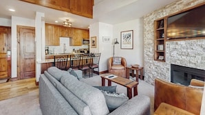 Ein Wohnzimmer mit Sofa, Couchtisch und Kamin. Dazu eine Küchenzeile mit Holzschränken und einem Esstisch.