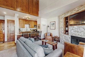 Ein Wohnzimmer mit Sofa, Couchtisch und Kamin. Dazu eine Küchenzeile mit Holzschränken und einem Esstisch.
