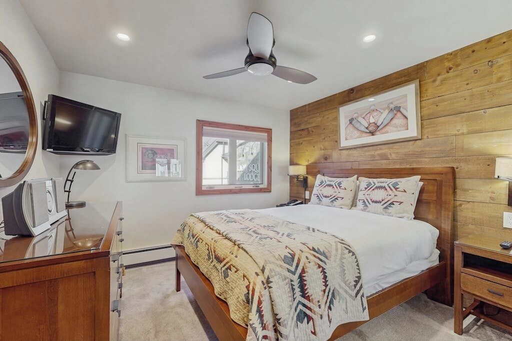 A bedroom with a bed, a ceiling fan, a TV, and wooden paneling on the wall.