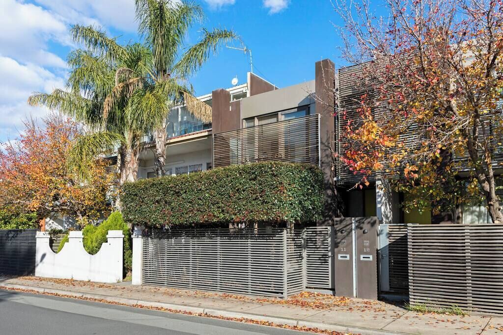 A modern two-story house with a flat roof, a large hedge, and a metal fence.