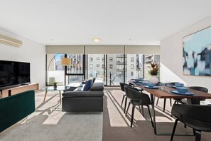 Ein modernes Wohnzimmer mit einer Couch, einem Fernseher, einem Essbereich mit Tisch und Stühlen sowie großen Fenstern mit Blick auf die Stadt.