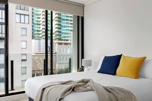 Ein Schlafzimmer mit einem großen Bett, einem Fenster mit Blick auf Gebäude und einem Nachttisch mit einer Lampe.