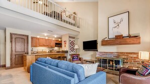 A spacious living area with a blue sectional sofa, a fireplace, and a kitchen in the background. - Ski-in/out Condo with fast WiFi, grill, fireplace & shared pool, hot tubs, W/D (Vail)
