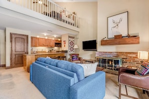 A spacious living area with a blue sectional sofa, a fireplace, and a kitchen in the background. - Ski-in/out Condo with fast WiFi, grill, fireplace & shared pool, hot tubs, W/D (Vail)