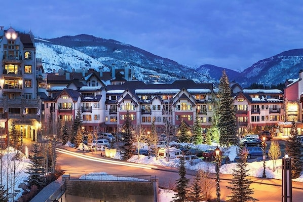 A hotel with multiple buildings, a parking lot, and snow-covered mountains in the background. - Ski-in/out Condo with fast WiFi, grill, fireplace & shared pool, hot tubs, W/D (Vail)