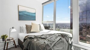 Un dormitorio con un ventanal grande, una cama con almohadas, una mesita de noche con lámpara y un cuadro en la pared.