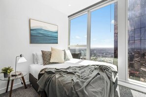 Ein Schlafzimmer mit großem Fenster, einem Bett mit Kissen, einem Nachttisch mit Lampe und einem Bild an der Wand.