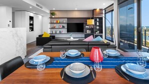 A modern living room with a black sofa, a dining table set for six, and a kitchen area with a TV and bookshelves. - Jaw-dropping Views from 3bdr Executive Penthouse (Melbourne)