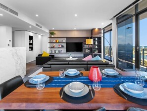 Ein modernes Wohnzimmer mit einem schwarzen Sofa, einem Esstisch für sechs Personen und einer Küchenzeile mit Fernseher und Bücherregalen.
