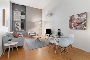 A modern living room with a grey sofa, a glass dining table with white chairs, a grey rug, a small side table with a plant, and a large window with a view of a brick wall. - Famous Degraves Lane Stunner with Parking! (Melbourne)