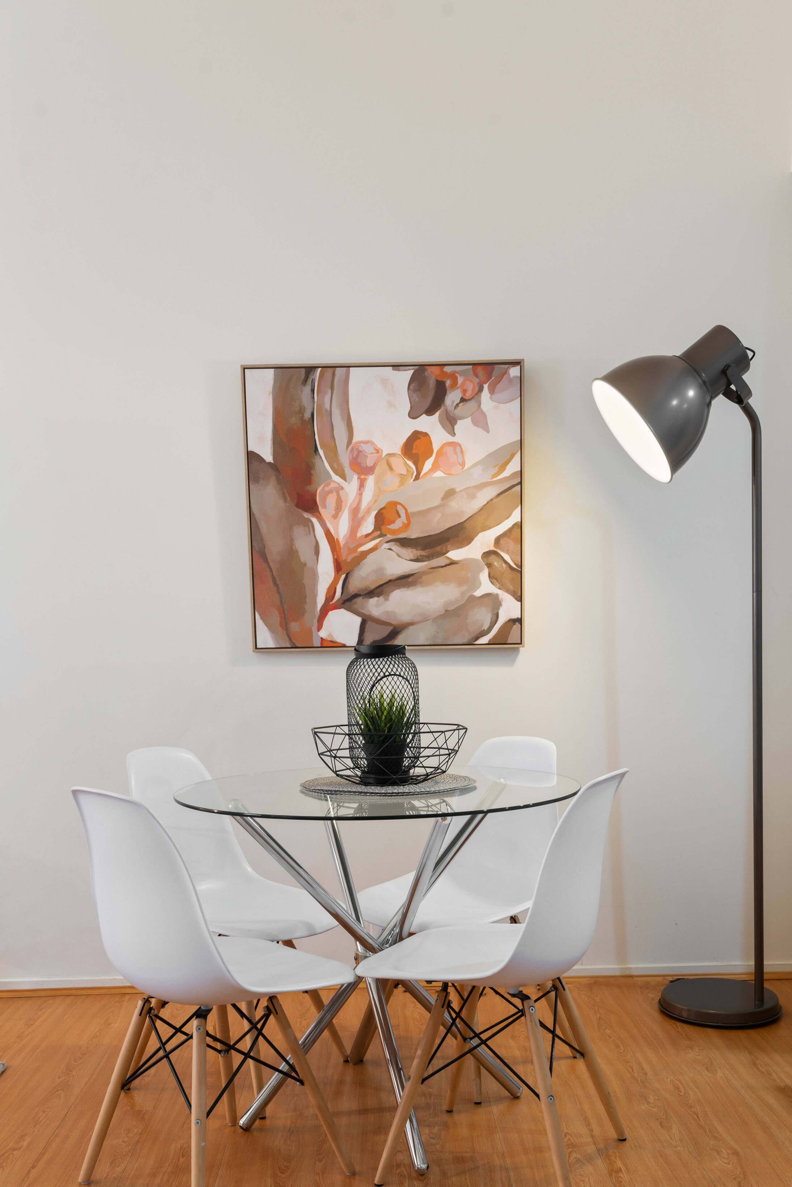 A dining area with a glass table, white chairs, a black floor lamp, and a framed abstract painting.