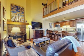 A spacious living area with a fireplace, a television, and a kitchen in the background. - Ski-in/out Luxury condo with fireplace, fast WiFi, shared pool/hot tub/laundry (Vail)