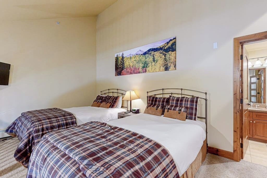 A hotel room with two beds, a nightstand with a lamp, a painting of a forest on the wall, and a wooden door.