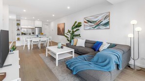Un salon moderne avec un canapé, une table basse et un téléviseur. On y trouve également un coin repas avec une table et des chaises. Une plante et une œuvre d’art sont visibles.