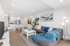 A modern living room with a sofa, coffee table, and a TV. There is a dining area with a table and chairs. A plant and artwork are visible. - Executive 3BD City Haven with Parking + Views (Melbourne)