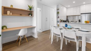Une cuisine moderne avec des meubles blancs, des étagères en bois et un coin repas avec des chaises blanches.