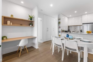 A modern kitchen with white cabinetry, wooden shelves, and a dining area with white chairs. - Executive 3BD City Haven with Parking + Views (Melbourne)