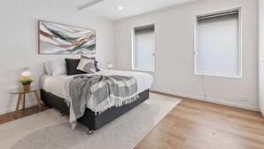 Ein modernes Schlafzimmer mit einem Bett, Nachttischen, einem an der Wand befestigten abstrakten Gemälde und zwei Fenstern.
