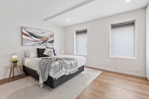 A modern bedroom with a bed, bedside tables, a wall-mounted abstract painting, and two windows.