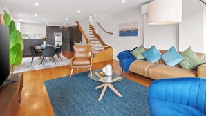 Ein modernes Wohnzimmer mit einem Glastisch, einer Couch, einem braunen Ledersessel, einer Essecke mit Tisch und Stühlen sowie einer Treppe.