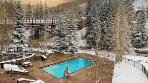 Un espace piscine doté d’une terrasse en bois, entouré d’arbres couverts de neige et avec une chaîne de montagnes en toile de fond.