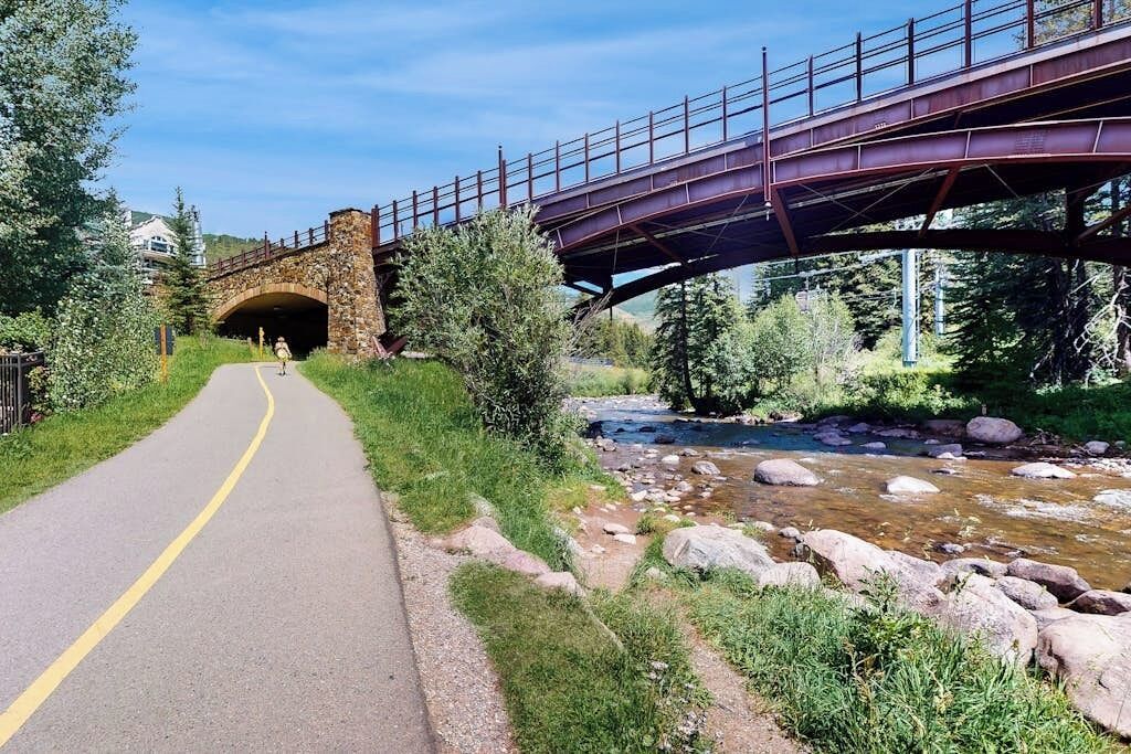 Un sendero pavimentado bajo un puente, con un arroyo a un lado.