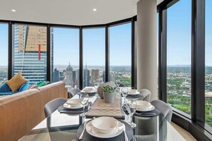 A dining area with a table set for four, large windows offering a city view, and a sofa with cushions. - Live in the clouds! 3BD luxury apt. in Southbank. (Southbank)