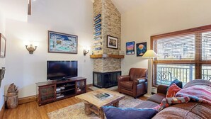 A living room with a fireplace, television, leather chair, and a large window. - Elegant, ski-in/out condo w/ fireplace, fast WiFi, shared pool/hot tubs/laundry (Vail)