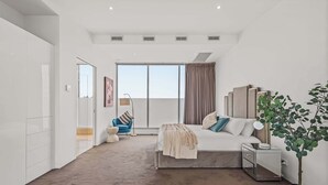 Quarto moderno com uma cama grande, mesas de cabeceira, um criado-mudo, uma planta em vaso, uma cadeira e vista para o exterior.