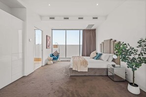 Ein modernes Schlafzimmer mit einem großen Bett, Nachttischen, einem Beistelltisch, einer Topfpflanze, einem Sessel und Blick ins Freie.